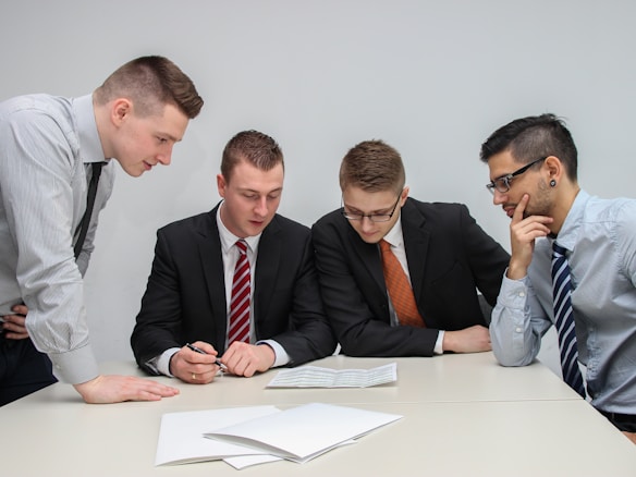 A group of four men in business attire are gathered around a table, examining documents laid out in front of them. They appear to be engaged in a discussion or meeting, with focused expressions and attentive body language.