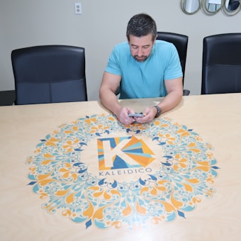 A man in a light blue shirt is seated at a large conference table, which has a colorful circular design featuring an intricate pattern and the word 'Kaleidico' in the center. The table is set in a modern office environment with black chairs, a wall with mirrors, and electronic equipment in the background.