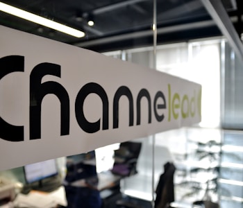 A modern office setting with a focus on a glass partition displaying the word 'Channel' printed in large black letters and 'lead' in lighter green letters. The background shows a blurred view of desks, papers, computers, office chairs, and vertical blinds, indicating an active work environment.