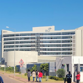 A modern, large, multi-story office building is prominently situated on a clear day, featuring the letters 'BCI' with a logo on its facade. The structure is surrounded by a well-maintained garden and a sidewalk where several people are walking. Nearby, cars are parked and some are driving along the street.